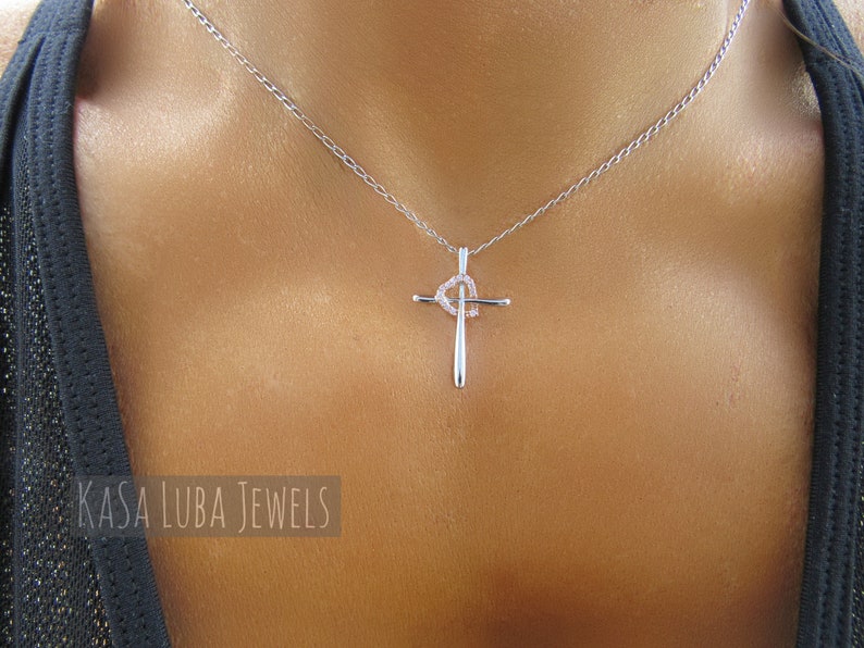 Puede incluir: Un collar de cadena de plata con un colgante de cruz de plata que presenta un peque&ntilde;o dise&ntilde;o de coraz&oacute;n con piedras transparentes. El collar lo lleva una persona con la piel bronceada.