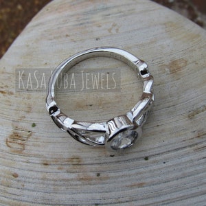 K&ouml;nnte beinhalten: Ein silberner Ring mit einem klaren, runden Edelstein. Der Ring hat ein dekoratives, blattartiges Design am Band. Der Text "KASA KUBA JEWELS" ist auf dem Ring zu sehen. Der Ring ist vor einem neutralen Hintergrund platziert.