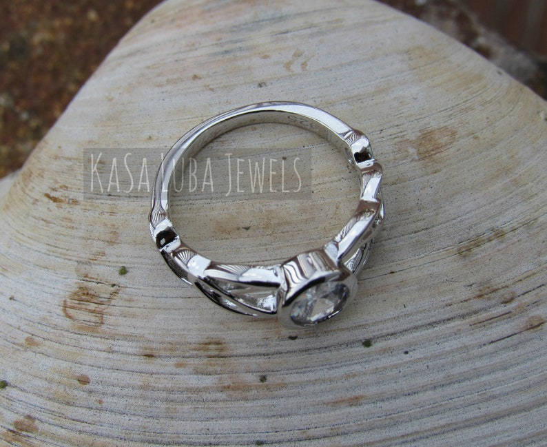 K&ouml;nnte beinhalten: Ein silberner Ring mit einem klaren, runden Edelstein. Der Ring hat ein dekoratives, blattartiges Design am Band. Der Text "KASA KUBA JEWELS" ist auf dem Ring zu sehen. Der Ring ist vor einem neutralen Hintergrund platziert.