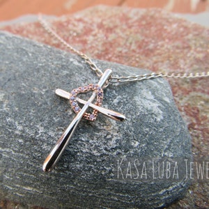Puede incluir: Un collar de cadena de plata con un colgante de cruz de oro rosa con un dise&ntilde;o de coraz&oacute;n con circonitas c&uacute;bicas transparentes. El colgante est&aacute; acentuado con un coraz&oacute;n de plata. El collar se muestra sobre una roca gris con el texto "Kasa Luba Jewels".