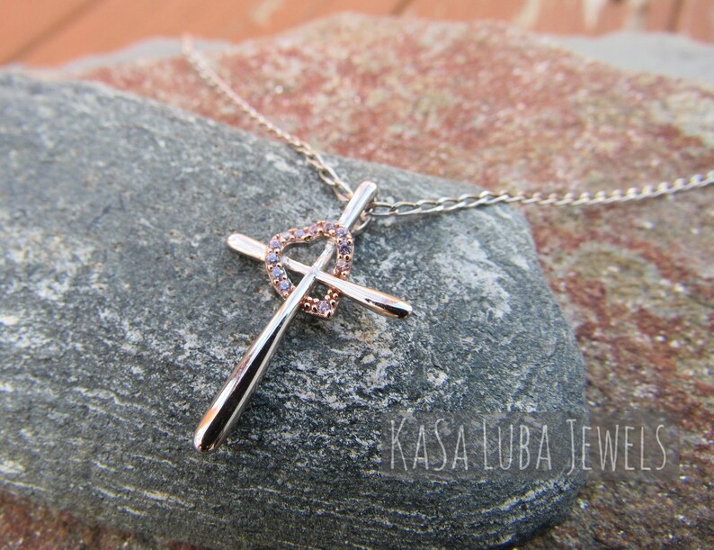 Puede incluir: Un collar de cadena de plata con un colgante de cruz de oro rosa con un dise&ntilde;o de coraz&oacute;n con circonitas c&uacute;bicas transparentes. El colgante est&aacute; acentuado con un coraz&oacute;n de plata. El collar se muestra sobre una roca gris con el texto "Kasa Luba Jewels".