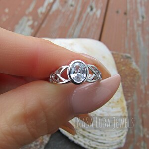 K&ouml;nnte beinhalten: Ein silberner Ring mit einem ovalen, klaren Edelstein. Der Ring hat ein keltisches Knotenmuster auf dem Band. Der Text "KASA LUBA JEWELS" ist sichtbar.
