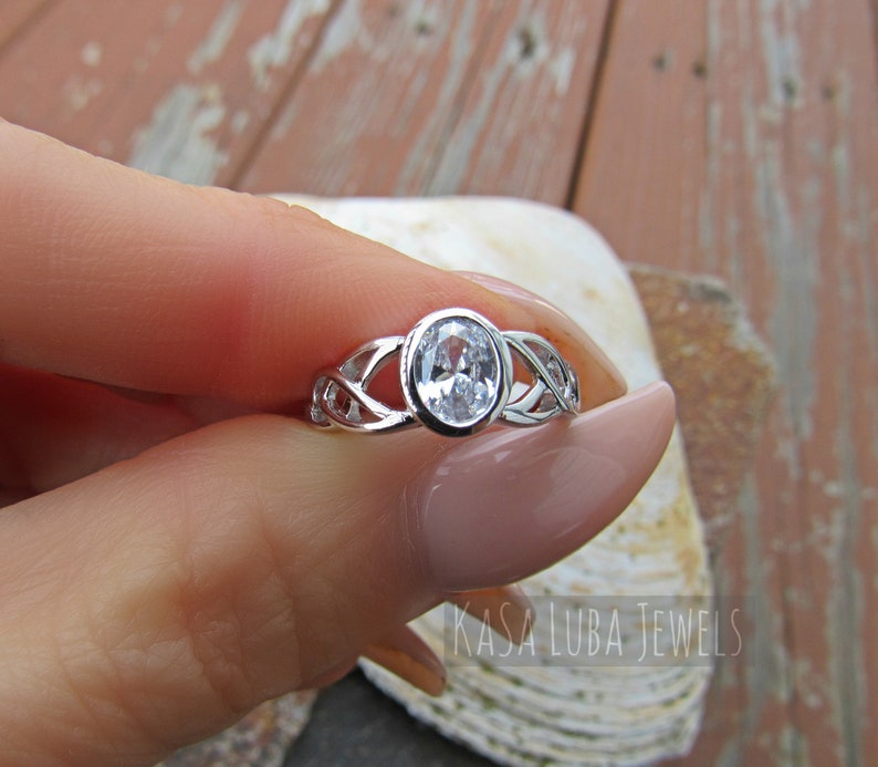 K&ouml;nnte beinhalten: Ein silberner Ring mit einem ovalen, klaren Edelstein. Der Ring hat ein keltisches Knotenmuster auf dem Band. Der Text "KASA LUBA JEWELS" ist sichtbar.