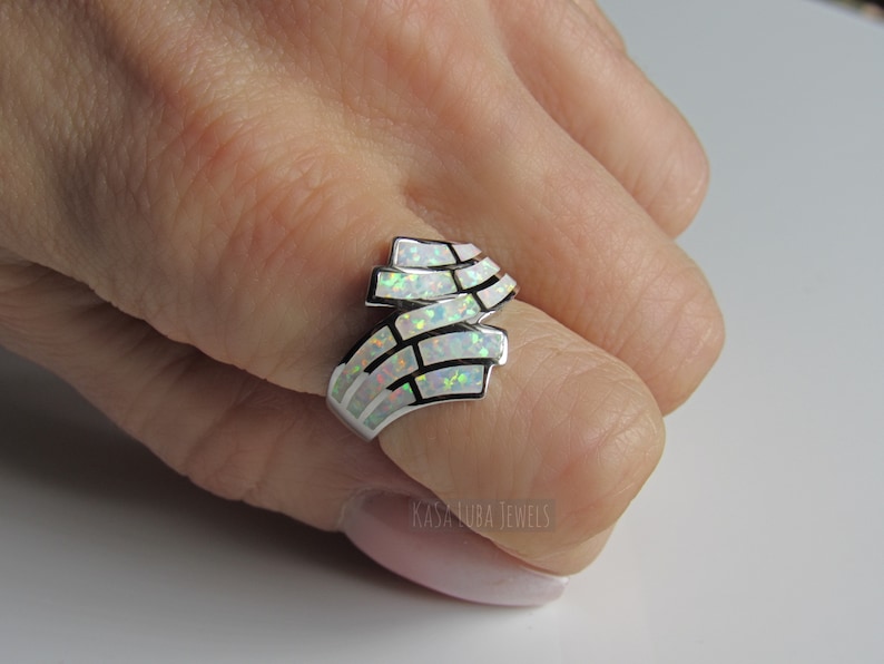 Op de afbeelding: Een zilveren ring met een witte opaal-steen gezet in een geometrisch ontwerp. De ring wordt gedragen aan een vinger.