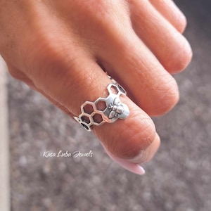 May include: A silver-colored ring featuring a honeycomb design with a bee accent. The ring's design incorporates hexagonal shapes, with a detailed bee at the center. The ring is worn on a finger, showcasing the intricate details of the jewelry.