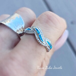 May include: A silver ring with a wavy design featuring blue opal stones and clear cubic zirconia stones. The ring is on a finger and the text "Kasa Luba Jewels" is visible below the ring.
