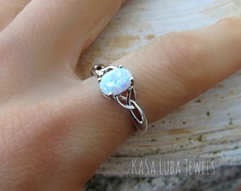 May include: A silver ring with a white opal gemstone set in a Celtic knot design. The ring is on a finger.