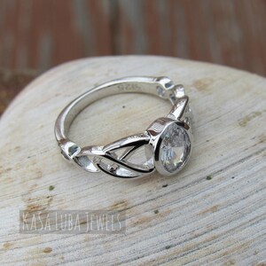 K&ouml;nnte beinhalten: Ein silberner Ring mit einem gro&szlig;en, klaren, oval geschliffenen Edelstein. Das Band hat ein kompliziertes keltisches Knotenmuster. Der Ring liegt auf einer hellen Oberfl&auml;che. Der Text "KASA LUBA JEWELS" ist sichtbar.