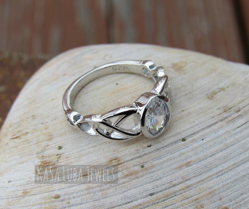 K&ouml;nnte beinhalten: Ein silberner Ring mit einem gro&szlig;en, klaren, oval geschliffenen Edelstein. Das Band hat ein kompliziertes keltisches Knotenmuster. Der Ring liegt auf einer hellen Oberfl&auml;che. Der Text "KASA LUBA JEWELS" ist sichtbar.