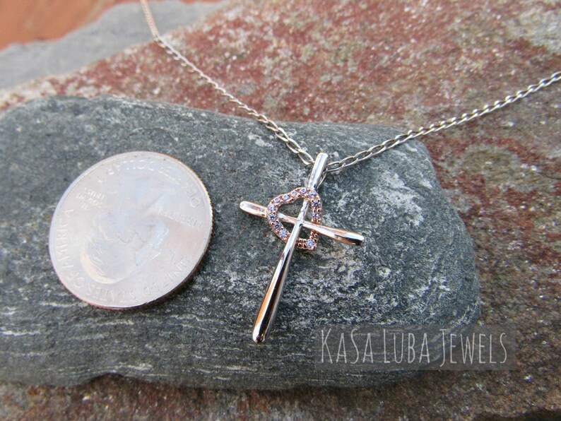 Puede incluir: Un collar de plata y oro rosa con un colgante de cruz con un dise&ntilde;o de coraz&oacute;n con circonitas c&uacute;bicas transparentes. El colgante es aproximadamente del tama&ntilde;o de una moneda de 25 centavos de d&oacute;lar estadounidense. El collar est&aacute; sobre una roca gris con el texto "KASA LUBA JEWELS" visible.