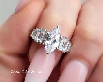 Könnte beinhalten: Ein silberner Ring mit einem großen, markisenförmigen Diamanten und kleineren, baguetteförmigen Diamanten auf der Ringschiene. Der Ring wird an einem Finger mit rosa und weißem Nagellack getragen.