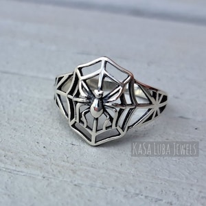 May include: A silver ring with a spider in a web design. The ring is on a light gray wooden surface. The text "Kasa Luba Jewels" is visible in the bottom right corner of the image.