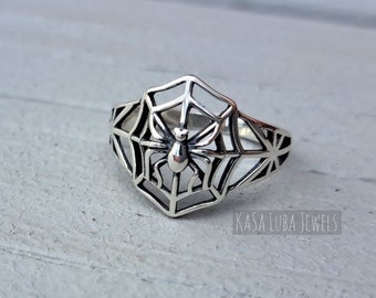 May include: A silver ring with a spider in a web design. The ring is on a light gray wooden surface. The text "Kasa Luba Jewels" is visible in the bottom right corner of the image.
