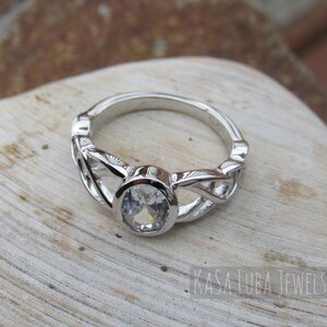 K&ouml;nnte beinhalten: Ein silberner Ring mit einem gro&szlig;en, klaren, runden Edelstein in einer L&uuml;nette. Das Band weist ein kompliziertes keltisches Knotendesign auf. Der Ring wird auf einer hellen, strukturierten Oberfl&auml;che pr&auml;sentiert.