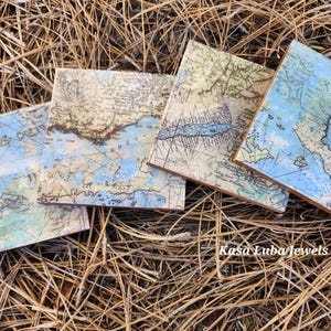 May include: Four square coasters featuring vintage nautical map prints in shades of blue, beige, and green. Each coaster displays detailed cartographic lines and geographical markings. The text Kasa Luba Jewels is printed in white on the bottom right.