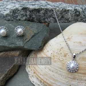 K&ouml;nnte beinhalten: Ein silbernes Halsketten- und Ohrring-Set mit Perlen- und Kristall-Akzenten. Die Halskette hat eine zarte Kette und einen Anh&auml;nger mit einer Perle, die von Kristallen umgeben ist. Die Ohrringe sind Ohrstecker mit einer Perle, die von Kristallen umgeben ist.