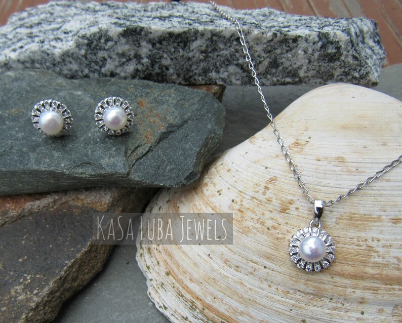 K&ouml;nnte beinhalten: Ein silbernes Halsketten- und Ohrring-Set mit Perlen- und Kristall-Akzenten. Die Halskette hat eine zarte Kette und einen Anh&auml;nger mit einer Perle, die von Kristallen umgeben ist. Die Ohrringe sind Ohrstecker mit einer Perle, die von Kristallen umgeben ist.