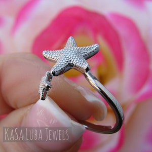 May include: Silver ring with a starfish design. The ring is open and has a smaller starfish charm hanging from it. The ring is shown against a pink background.