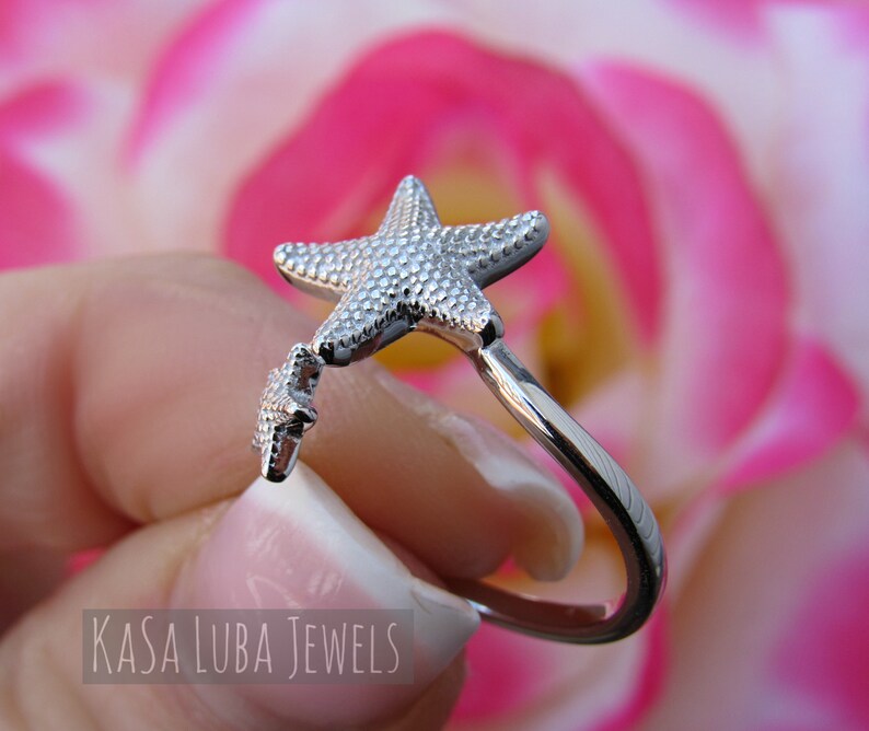 May include: Silver ring with a starfish design. The ring is open and has a smaller starfish charm hanging from it. The ring is shown against a pink background.