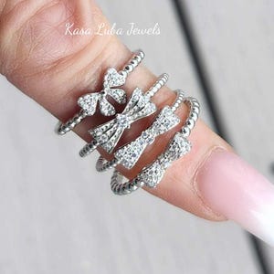 May include: A set of four silver rings with a bow design and clear cubic zirconia stones. The rings are stacked on a finger.