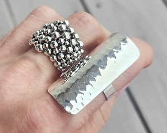 May include: Two silver rings are displayed. One ring features a cluster of small, round beads, while the other is a large, rectangular band with a hammered texture. The rings are on a hand, showcasing the jewelry's design and style.
