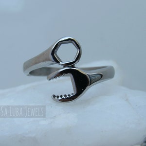 May include: A silver ring shaped like a wrench. The ring is open on one side and has a hexagonal shape on the other side. The ring is made of metal and has a shiny finish.