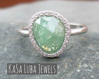 Puede incluir: Un anillo de plata con una gran piedra preciosa verde facetada rodeada por un halo de piedras blancas más pequeñas. El anillo está sobre una superficie gris texturizada. El texto "KASA LUBA JEWELS" es visible debajo del anillo.