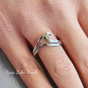 May include: A silver ring with a wave design. The ring is on a person's finger. The text "Kasa Luba Jewels" is visible in the image.
