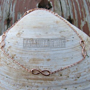Puede incluir: Pulsera de oro rosa con tres símbolos de infinito en una cadena. La pulsera está sobre un fondo de concha blanca. "Kasa Luba Jewels" está escrito en negro en la concha.