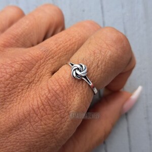 May include: A silver knot ring with a simple design. The ring is worn on a finger and is a delicate piece of jewelry.