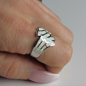 Op de afbeelding: Een zilveren ring met een witte opaal-steen gezet in een geometrisch ontwerp. De ring is op een hand, en de tekst "Kasa Luba Jewels" is zichtbaar in de linkerbenedenhoek van de afbeelding.