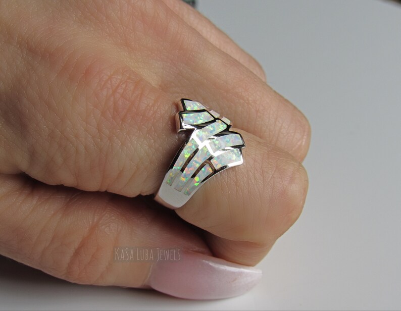 Op de afbeelding: Een zilveren ring met een witte opaal-steen gezet in een geometrisch ontwerp. De ring is op een hand, en de tekst "Kasa Luba Jewels" is zichtbaar in de linkerbenedenhoek van de afbeelding.