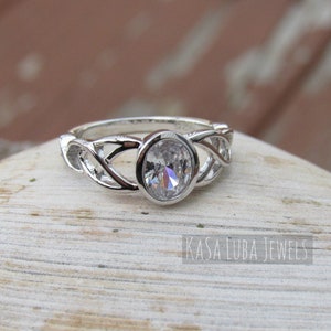 K&ouml;nnte beinhalten: Ein silberner Ring mit einem ovalen, klaren Edelstein in der Mitte. Das Band weist ein kompliziertes, ineinander verwobenes Design auf. Der Ring ist vor einem neutralen Hintergrund platziert. Der Text "KASA LUBA JEWELS" ist sichtbar.