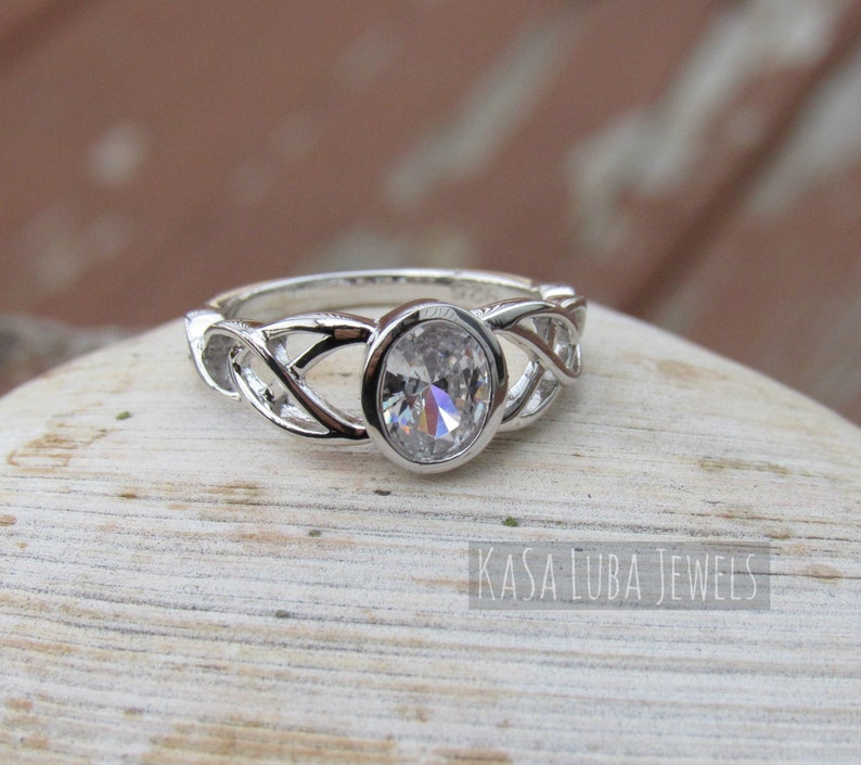 K&ouml;nnte beinhalten: Ein silberner Ring mit einem ovalen, klaren Edelstein in der Mitte. Das Band weist ein kompliziertes, ineinander verwobenes Design auf. Der Ring ist vor einem neutralen Hintergrund platziert. Der Text "KASA LUBA JEWELS" ist sichtbar.