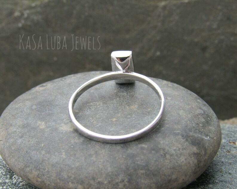 May include: A silver ring with a square-shaped gemstone. The ring is sitting on a gray rock.