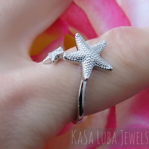 May include: Silver ring with two starfish designs. The larger starfish is on the top of the ring and the smaller starfish is on the side of the ring. The ring is on a finger.