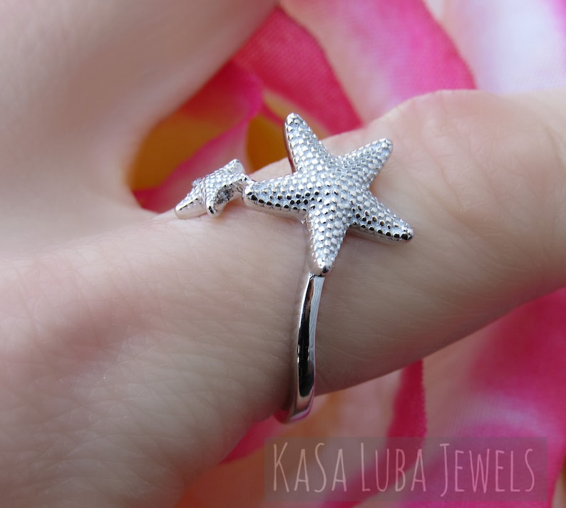 May include: Silver ring with two starfish designs. The larger starfish is on the top of the ring and the smaller starfish is on the side of the ring. The ring is on a finger.