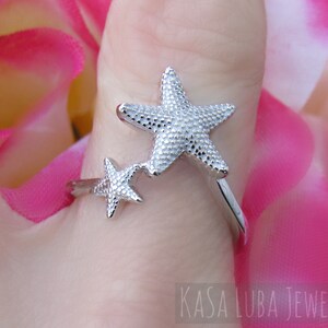May include: Silver ring with two starfish designs. The larger starfish is on the top and the smaller starfish is on the bottom. The ring is on a finger.