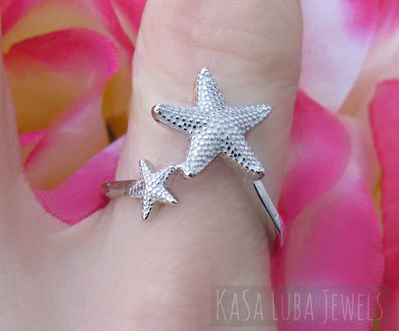 May include: Silver ring with two starfish designs. The larger starfish is on the top and the smaller starfish is on the bottom. The ring is on a finger.