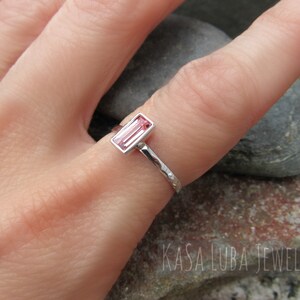 May include: A silver ring with a pink rectangular gemstone set in a bezel setting. The ring band is textured and has a hammered finish. The ring is worn on a finger.