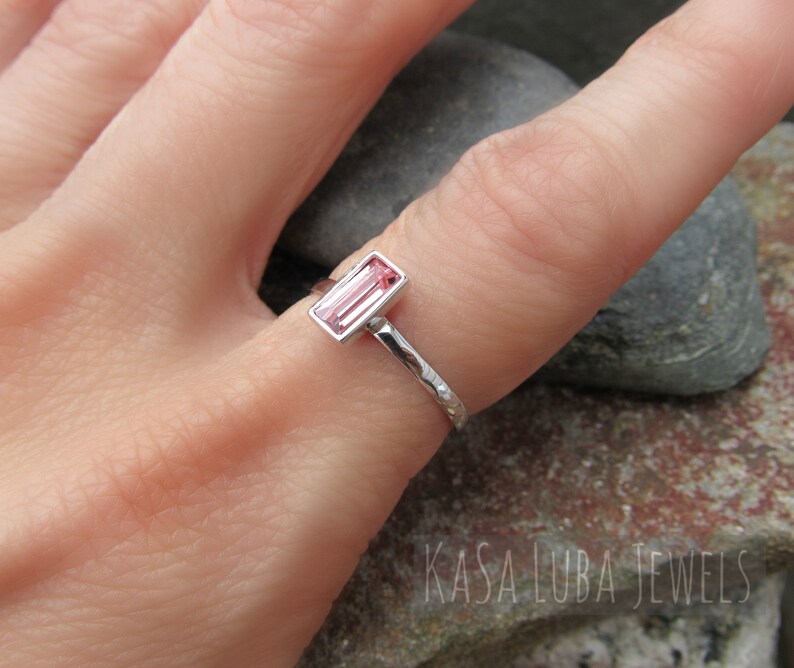 May include: A silver ring with a pink rectangular gemstone set in a bezel setting. The ring band is textured and has a hammered finish. The ring is worn on a finger.