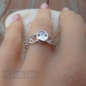 K&ouml;nnte beinhalten: Ein silberner Ring mit einem klaren, ovalen Edelstein. Der Ring hat ein kompliziertes keltisches Knotenmuster auf dem Band. Der Text "KASA LUBA JEWELS" ist im Bild zu sehen.