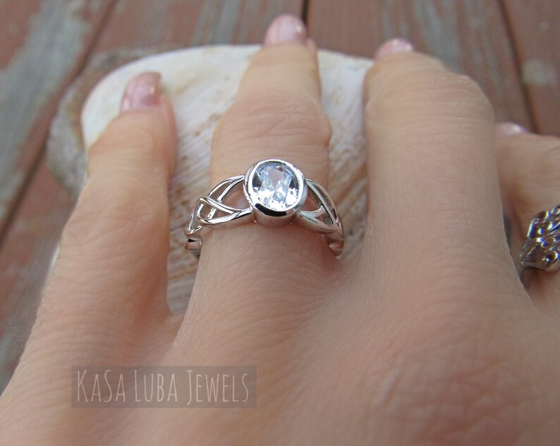 K&ouml;nnte beinhalten: Ein silberner Ring mit einem klaren, ovalen Edelstein. Der Ring hat ein kompliziertes keltisches Knotenmuster auf dem Band. Der Text "KASA LUBA JEWELS" ist im Bild zu sehen.