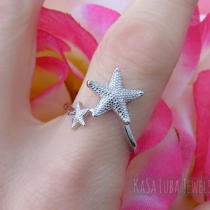 May include: Silver ring with two starfish designs. The larger starfish is textured and the smaller starfish is smooth. The ring is on a finger wearing a pink flower.