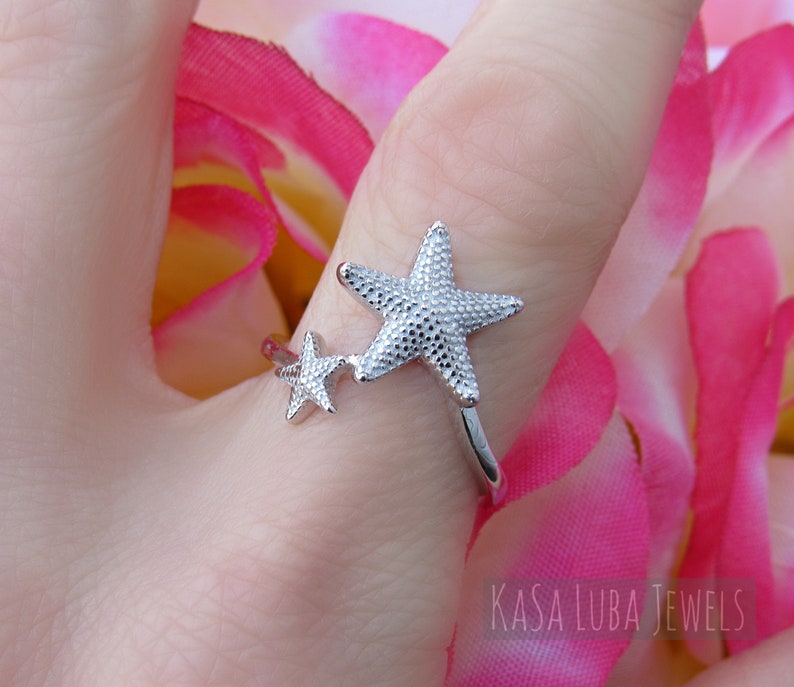 May include: Silver ring with two starfish designs. The larger starfish is textured and the smaller starfish is smooth. The ring is on a finger wearing a pink flower.