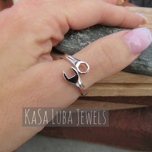 May include: A silver adjustable ring featuring two wrench designs. The ring is worn on a person's thumb.