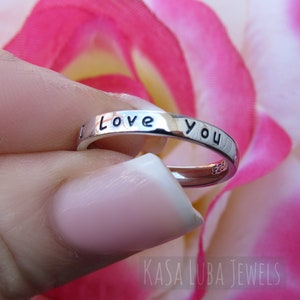 May include: A silver ring with the words "I love you" engraved on the outside. The ring is held against a pink and white floral background. The ring is marked with "925".