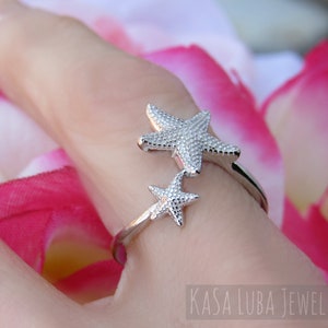 May include: Silver ring with two starfish designs. The ring is on a finger and the background is pink and white.
