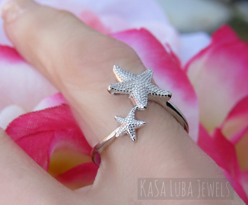 May include: Silver ring with two starfish designs. The ring is on a finger and the background is pink and white.