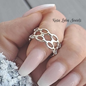 May include: A silver chain ring with a delicate, interlocking design. The ring is worn on a finger with a light pink ombre nail polish.